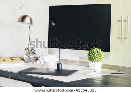 Sideview of designer desktop with black computer screen, graphic tablet, alarm clock and other items. Concrete wall and glass door in the background. Mock up