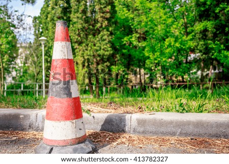 A road cone standing next to sidewalk's edge with trees in background near summertime