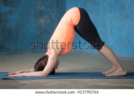 Beautiful young woman dressed in bright sportswear enjoying yoga indoors. Yogi girl working out in grunge interior with blue wall. Stretching in Dolphin Pose. Full length