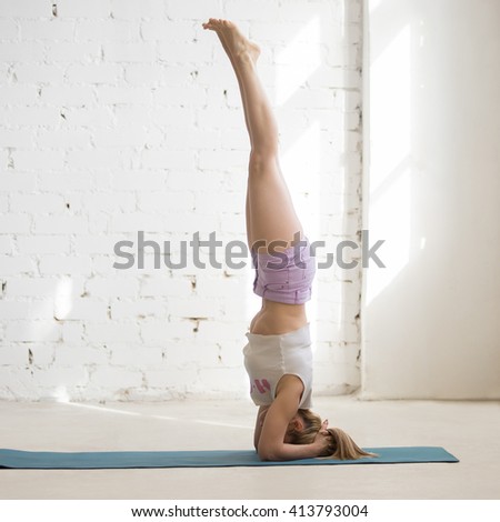 Beautiful young woman wearing casual clothing doing yoga indoors. Yogi girl working out in sunny loft interior. Doing supported headstand, salamba sirsasana. Square image. Full length. Side view