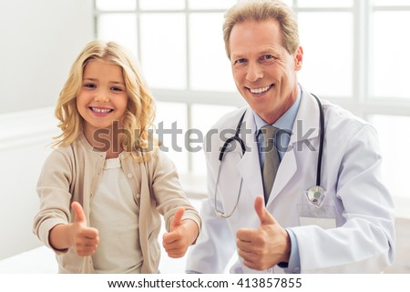 Cute little girl and handsome middle aged pediatrician are showing Ok sign, looking at camera and smiling, sitting in doctors office