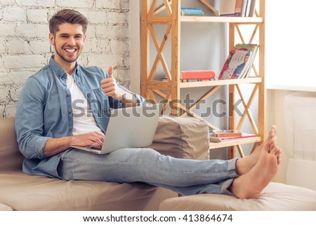 Attractive young man is using a laptop, showing Ok sign, looking at camera and smiling while sitting on sofa at home