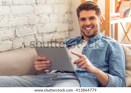 Attractive young man is using a tablet, looking at camera and smiling while lying on sofa at home