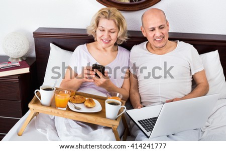 Mature positive couple reading news from notebook as having breakfast 