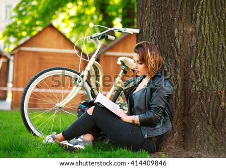 Young woman with bicycle reading a book in a city park on the grass