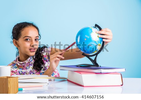Education at home concept - Cute little Indian/Asian girl studying or completing home work on study table with pile of books, educational globe, laptop computer, coffee mug etc