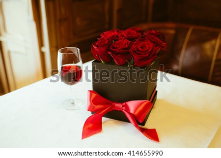 Beautiful red roses in a black box with a red ribbon with a glass of wine next on a table