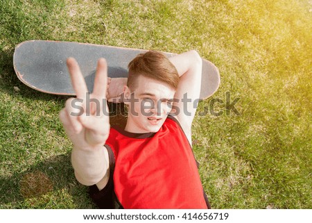 Young man lying on grass in park shows peace sign, skateboard near, summertime