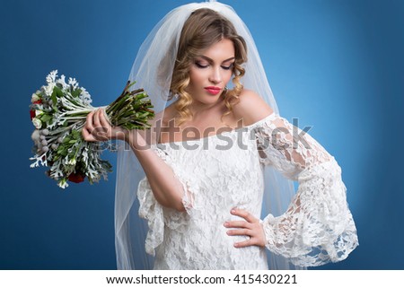 young beautiful girl in a wedding dress with a veil with a bouquet of flowers in hands on blue background