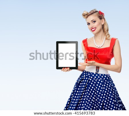 woman, showing blank no-name tablet pc monitor, with copyspace, dressed in pin-up style dress in polka dot, on blue background. Caucasian blond model posing in retro fashion vintage shoot.
