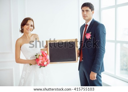 Wedding couple in love. Beautiful bride in white dress with brides bouquet and handsome groom in blue suite standing and holding impty board, sign for text indoors in decorated studio room, white