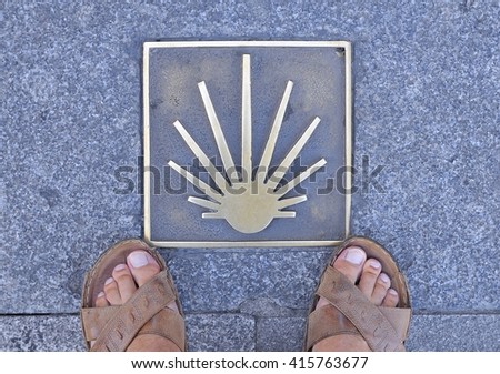 Metal signal in asphalt, Way of St. James in Spain, man's feet in sandals on sidewalk.From above