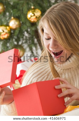 Beautiful teenage girl opening her christmas present. Christmas tree with golden glass balls in background
