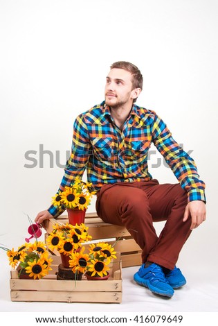 Young florist on a white background. Handsome young man in bright clothes sits on a wooden box with sunflowers in their hands. Care for flowers