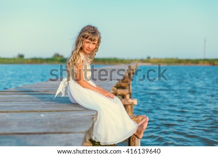 Little girl with long blond hair sitting on the pier near the sea