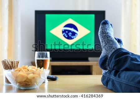 man watching Brazilian (Brazil) flag on TV screen with legs on table - stock photo