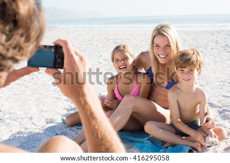 Father clicking picture of family at beach. Family posing for a photo during summer vacation. Cheerful mother sitting with son and daughter at beach for photo.
