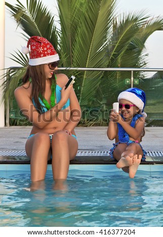 A funny woman with a child in Christmas hats, sitting poolside, take pictures on mobile phones