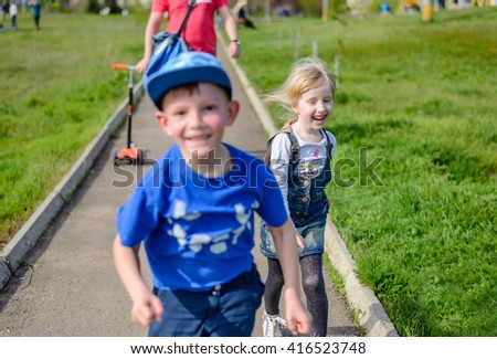 Vivacious spontaneous pretty little blond girl running and laughing along a path with her brother as they go out to play in the sunshine, fathers legs visible behind