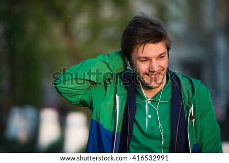Young man walking around city listening to music with ear headphones looks at the camera and smiling. Close-up shot. Sunset. Autumn weather.