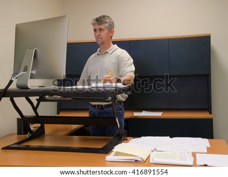 A man is working at a stand up desk in an office where he works because standing is healthier than sitting all day. Live healthy, do not sit all day. Royalty-Free Stock Photo #416891554