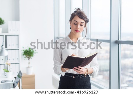 Female office worker writing down notes and day plan in her notebook, standing at workplace, front view portrait.