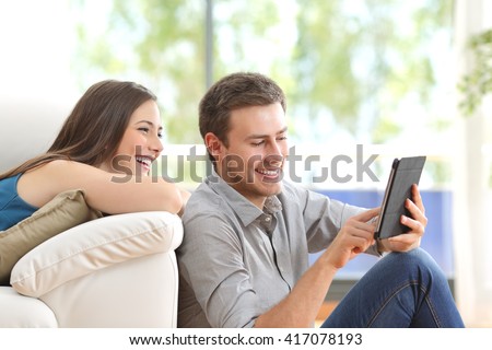 Cheerful couple using a tablet on line sitting in the living room at home with a window in the background