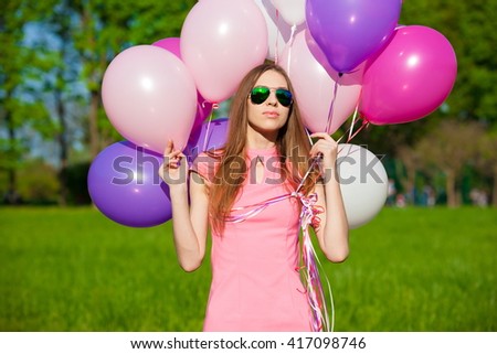 Beautiful fun woman holding multicolored helium balloons. Has smiling face, long hair, clothed pink dress, sunglasses. Has slim body. Portrait in green forest. Sunny day and blue sky. Close up.