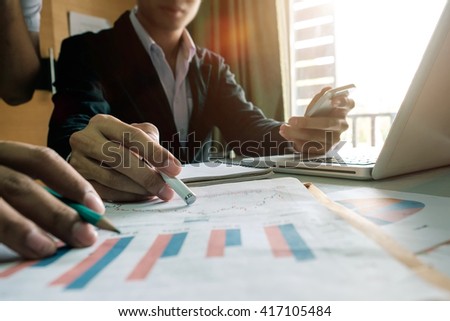 business documents on office table with smart phone and laptop and two colleagues discussing data in the background in morning light