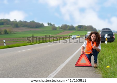 Smiling attractive woman putting out a traffic warning sign on the shoulder of the highway after breaking down in her car and giving thumb up gesture