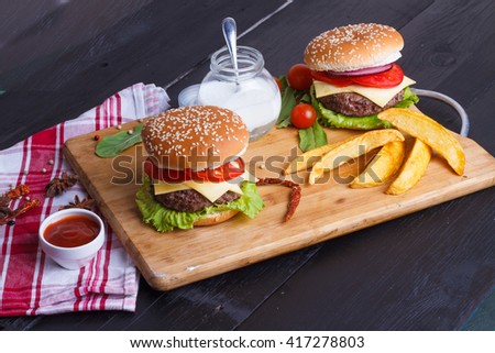 two burger sandwiches with potato wedges served on a wooden tray on a dark background