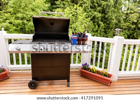 Open large barbecue cooker, bottled beer, cup, and crate on cedar wood deck with trees in background.  Royalty-Free Stock Photo #417291091