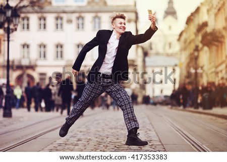Full body portrait of young happy smiling handsome man running, jumping & taking selfie photo on the old street. City lifestyle. Old european architecture on background. Toned style instagram filters
