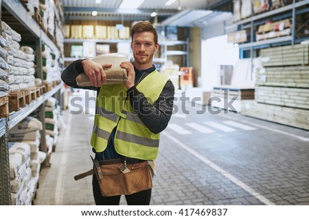 Young handyman selecting a pocket of product in a warehouse standing with the bag over his shoulder smiling at the camera Royalty-Free Stock Photo #417469837