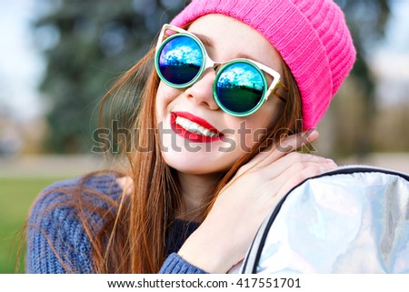 Fashion close up portrait of pretty ginger girl holding silver backpack and smiling, unusual trendy sunglasses, hipster neon hat, posing at city park.