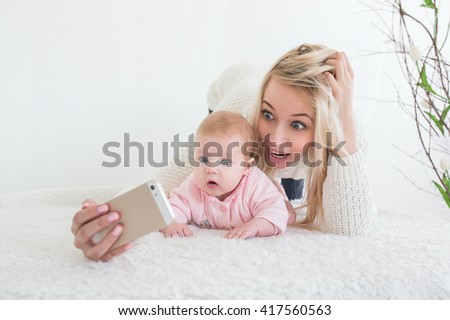 Funny baby girl make selfie on mobile phone and lying near her mother. Newborn looking at the camera and smiling. 