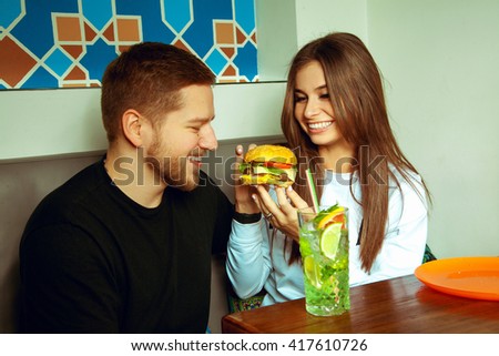 girl feeding her boyfriend hamburger. Couple in love having some fun in cafe
