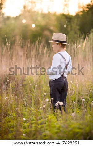 Adorable kid boy holding straw hat and standing on a summer meadow. Child walking in the park on a sunny summer day. Sunset in the park. Outdoors. Vacations.