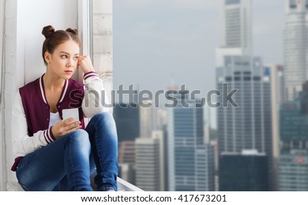 people, emotion, technology and teens concept - sad unhappy pretty teenage girl sitting on windowsill with smartphone and looking through window over singapore city skyscrapers background