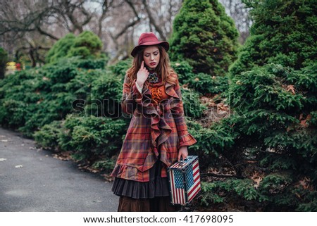 Beautiful girl in a red hat and stylish dress with a suitcase with the American flag stands in the park on a background of trees and shrubs. English lady. Alone retro concept