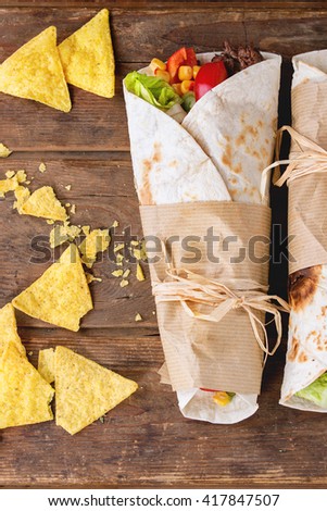 Mexican style. dinner. Two papered tortillas burrito with beef and vegetables served with nachos chips sauce over old wooden background. Flat lay