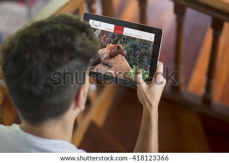 Young man sitting on the stairs at home using a tablet with online travel agency on screen. All screen graphics are made up.
