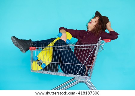 Fashion smiling hipster woman having fun wearing a hat with yellow skateboard sitting in the shopping trolley cart isolated over blue background