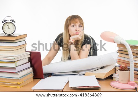 Girl student sitting sadly at the table crammed with books and papers