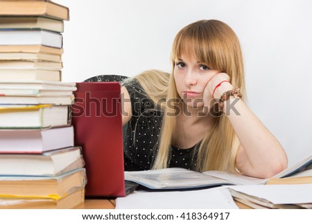 Girl student sitting at a desk with a laptop and sadly looks out from behind a pile of books