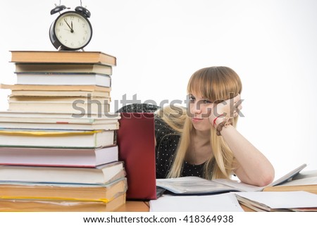 Sad girl student sitting at a desk with a large stack of books and looking at the laptop