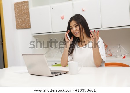 Portrait of happy Asian student communicating over mobile or smart phone and looking at laptop computer. Brunette in library.