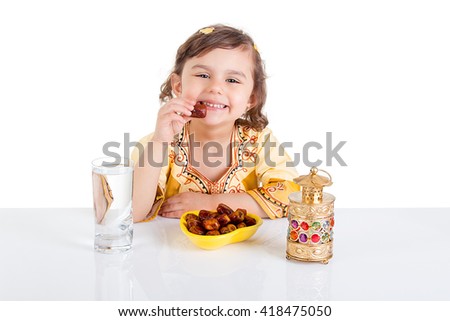 Young Muslim girl eating dates for brakfast in Ramadan - sitting on a table with water , dates , Quran, and Ramadan lantern Royalty-Free Stock Photo #418475050