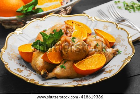 Baked chicken drumstick with baked oranges, fresh oranges cut for chicken, green mint, thyme, silver fork, a white porcelain plate, cloth, wicker basket on a dark black wooden rustic background