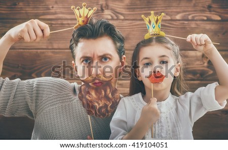 Funny family! Father and his child daughter girl with a paper accessories. Beauty funny girl holding paper lips and crown on stick. Beautiful young man holding paper beard and crown on stick. Royalty-Free Stock Photo #419104051